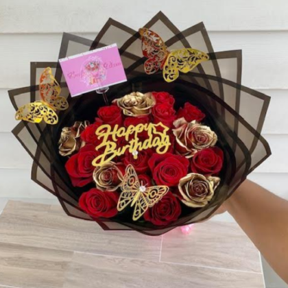 a stunning hand-tied bouquet featuring a harmonious blend of vibrant red roses and elegant gold roses. The rich red roses symbolize deep love and passion, while the gold roses add a touch of luxury and celebration. Interwoven throughout the bouquet are delicate gold butterflies, adding a whimsical and enchanting touch. At the center, a gold “Happy Birthday” emblem stands out, making the bouquet perfect for a special birthday celebration. The bouquet is finished with a lovely bow, tying everything together beautifully. The bow, in a matching gold or perhaps a contrasting color like deep red, adds the final touch of elegance and charm. This bouquet is not just a gift; it’s a statement of love, joy, and celebration, perfect for making someone’s birthday truly memorable. 🌹🎂✨