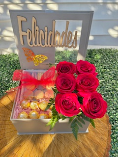 Deluxe gray gift box that includes vibrant red roses, fresh greens, in a beautiful arrangement with a chocolate gift surprise and delicious macarons that fill up a big portion of the box. Wood Emblem that reads Felicidades and a Butterfly that adds a special touch of elegance to this lovely gift.